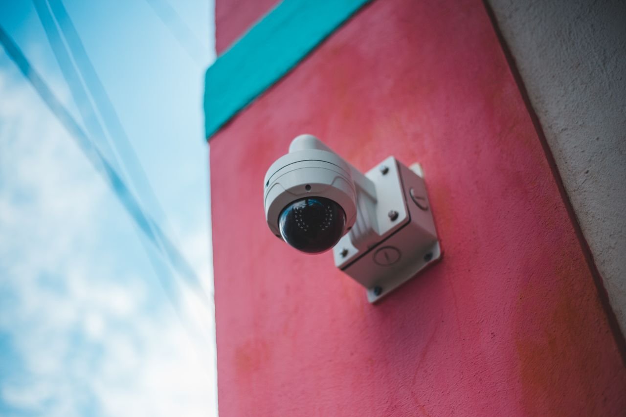 A surveillance camera mounted on a vibrant pink and teal wall outdoors.